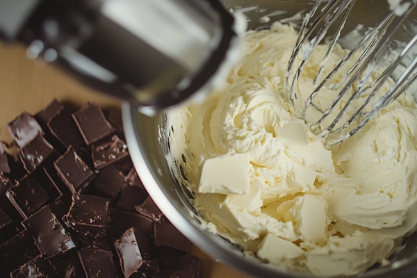 Whipped cream cheese and butter in a bowl for chocolate chip cheesecake cookies with chocolate pieces nearby.